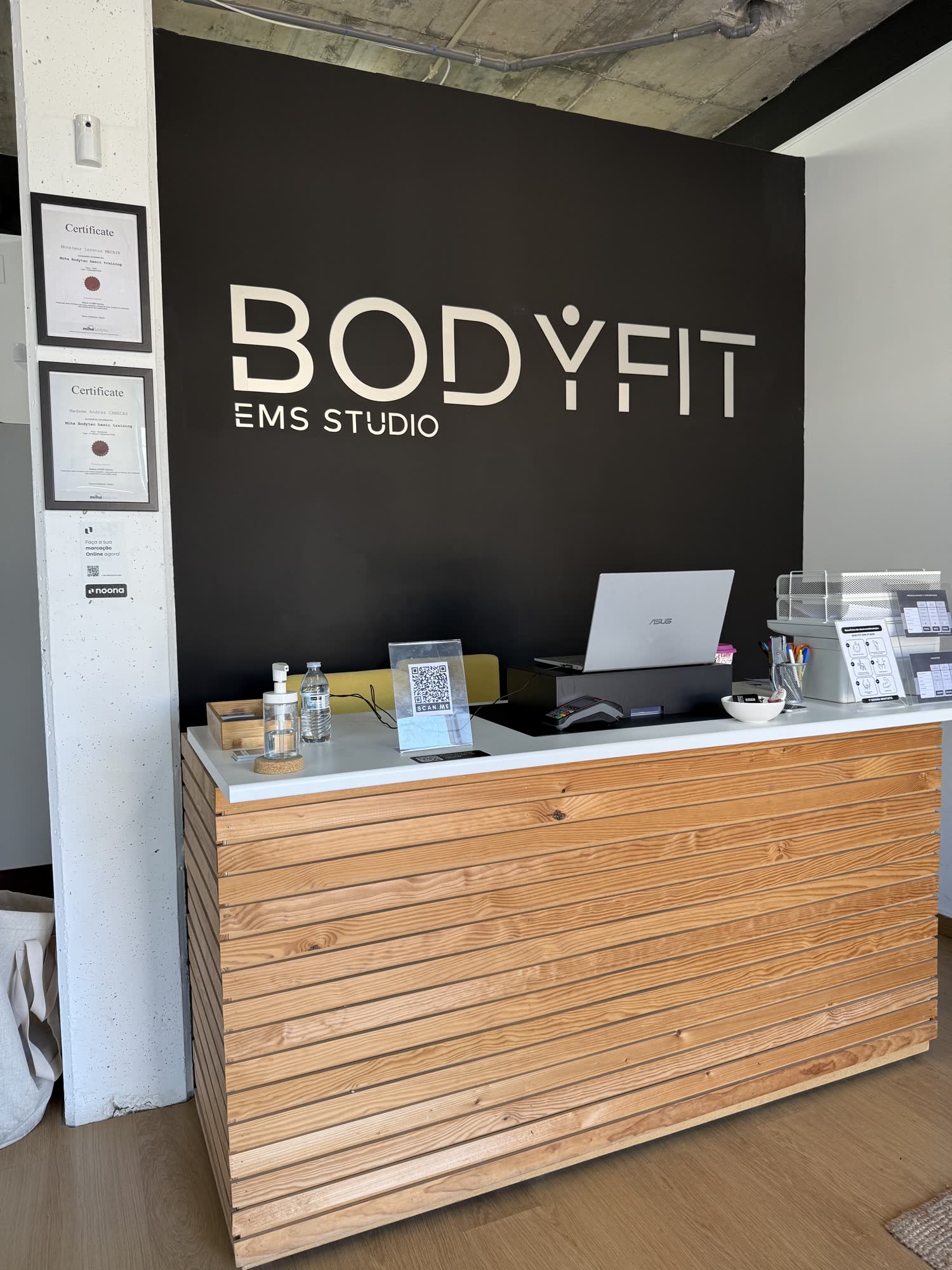 Receção do BODYFIT EMS Studio em Campo de Ourique, com o logo BODYFIT sobre parede escura, balcão em madeira e certificados na entrada.